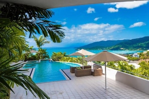 A pool area with a view of the ocean and hills, surrounded by palm trees and outdoor furniture.