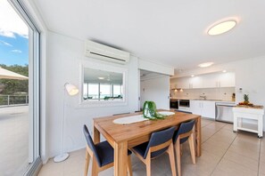 A modern kitchen with white cabinets, a wooden dining table, and a view through a large window. - Ocean Dreams Whitsunday (Airlie Beach)