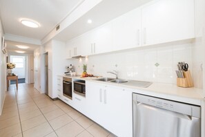 A modern kitchen with white cabinets, a stainless steel dishwasher, and a countertop with a fruit bowl and utensils. - Ocean Dreams Whitsunday (Airlie Beach)