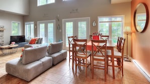 A spacious living room with a large sofa, dining table, and chairs. There are multiple windows providing natural light, and a television is visible in the background.