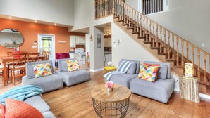 A spacious living room with a wooden staircase, grey sofas with colorful pillows, and a round coffee table with flowers.