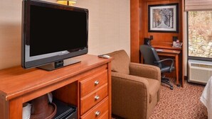 A hotel room with a television on a wooden desk, a brown armchair, a desk with a computer, and a framed picture on the wall. - McKnight Hotel (Pittsburgh)