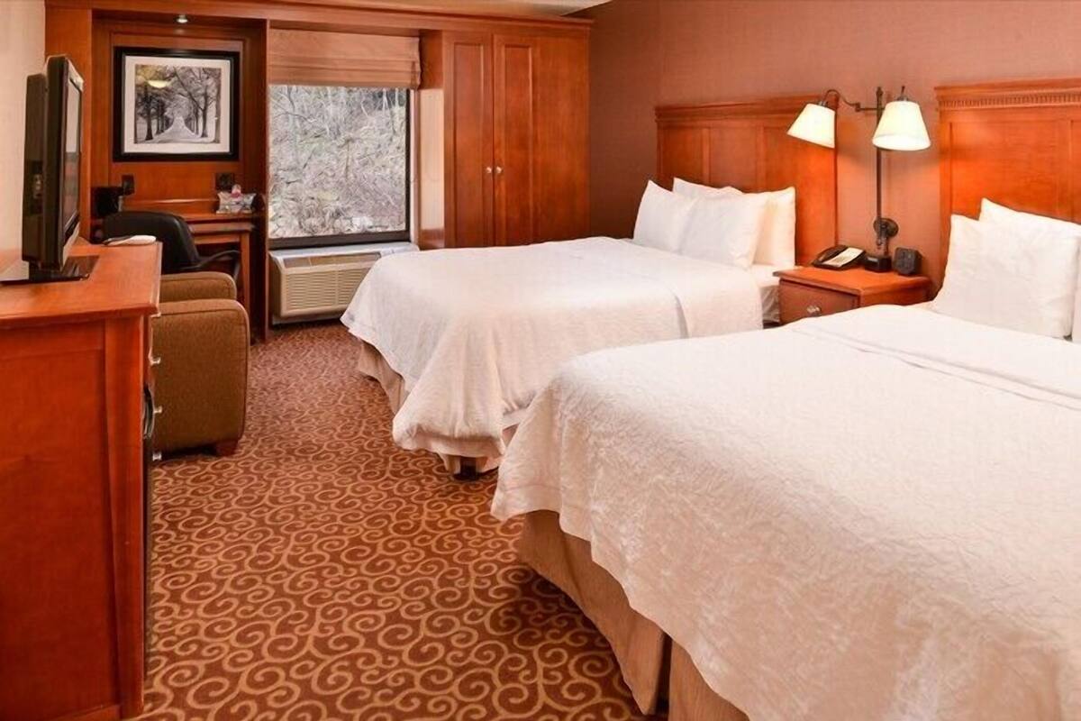 Ein Hotelzimmer mit zwei Betten, einem Schreibtisch, einem Stuhl und einem Fenster mit Blick auf Bäume.