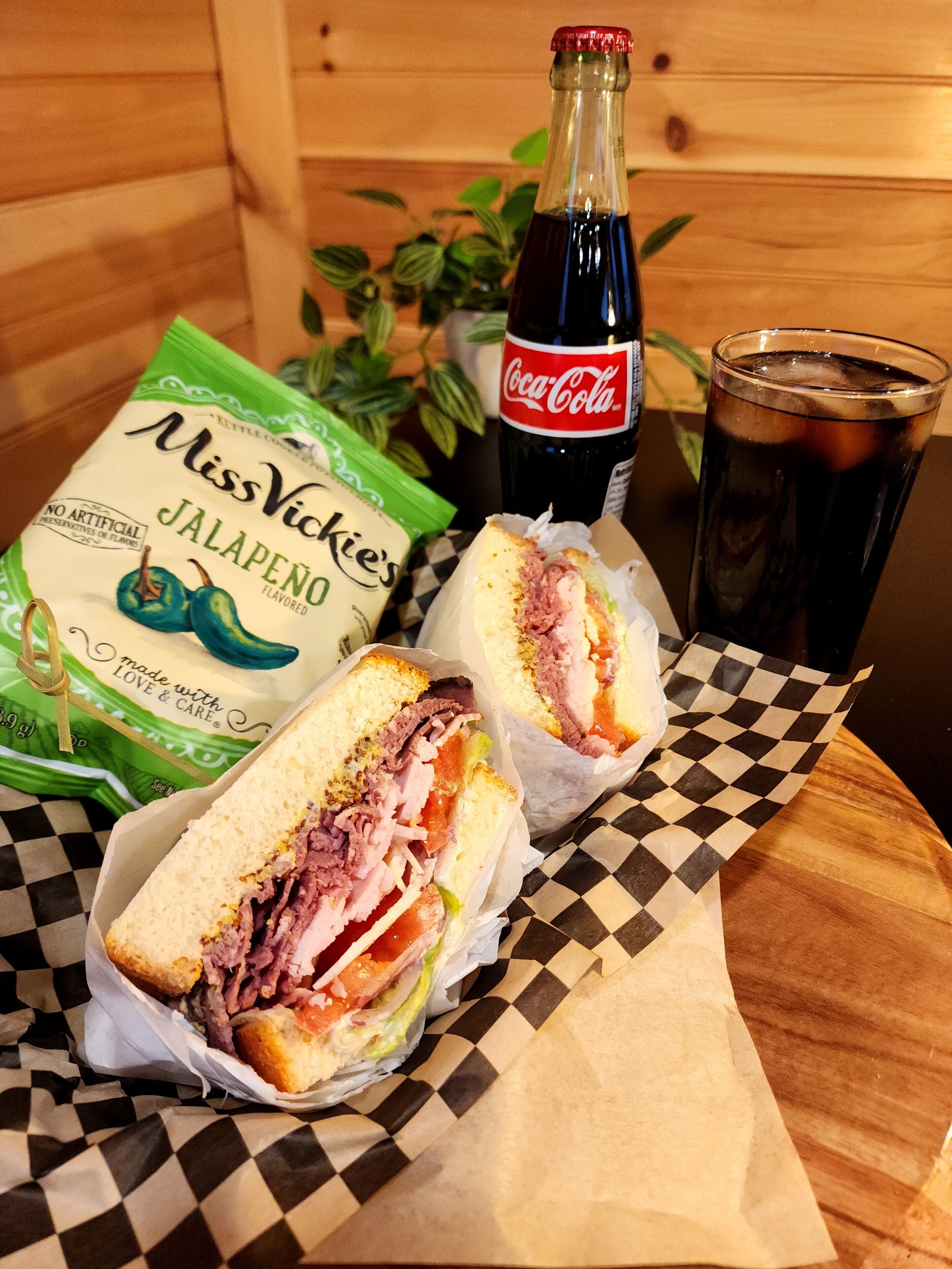 A sandwich, a bottle of Coca-Cola, and a bag of jalapeño-flavored chips on a checkered table.