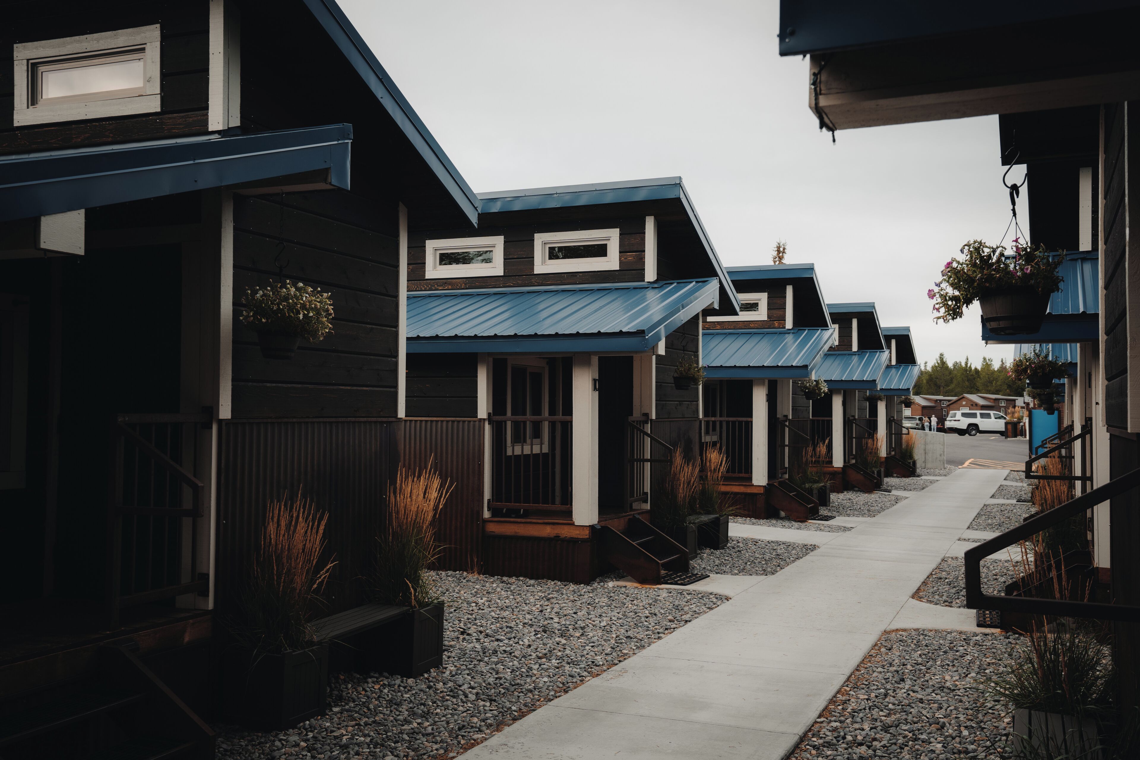 A row of modern, two-story buildings with blue roofs and wooden siding, arranged in a narrow street with gravel pathways.