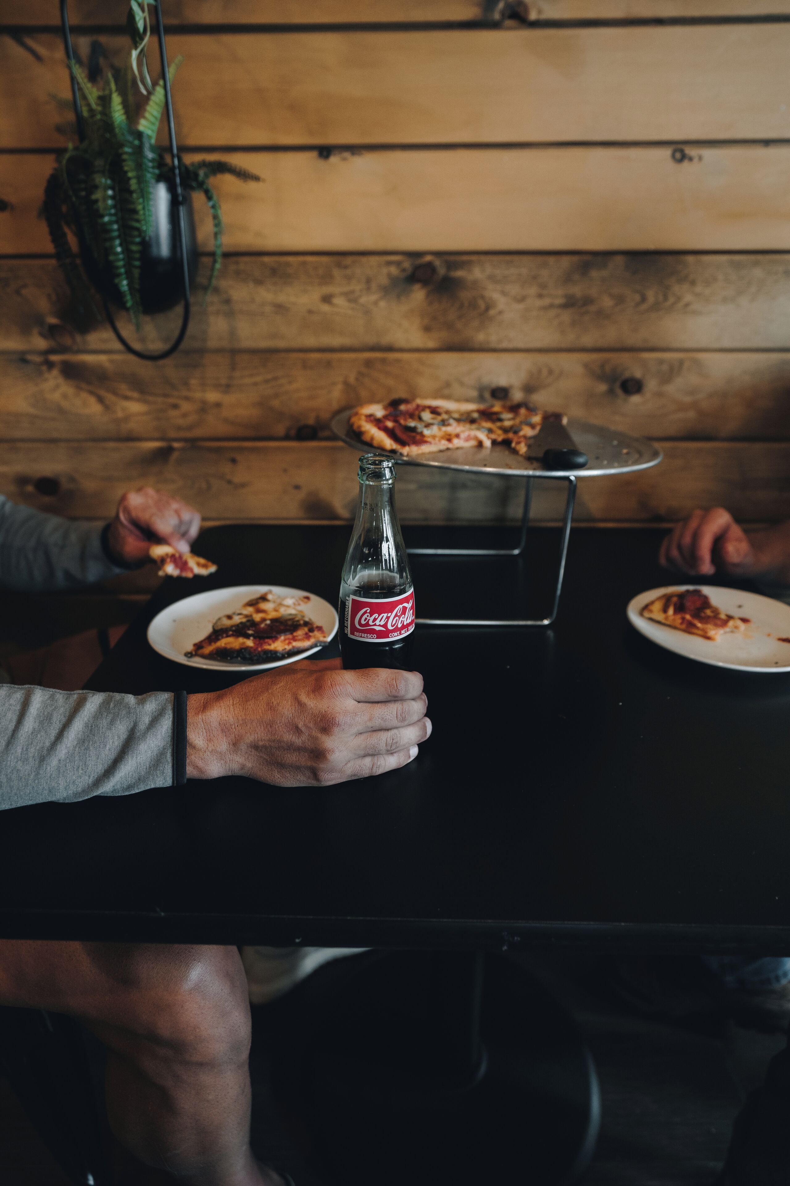 A wooden table with a Coca-Cola bottle, pizza slices, and a metal pizza stand.