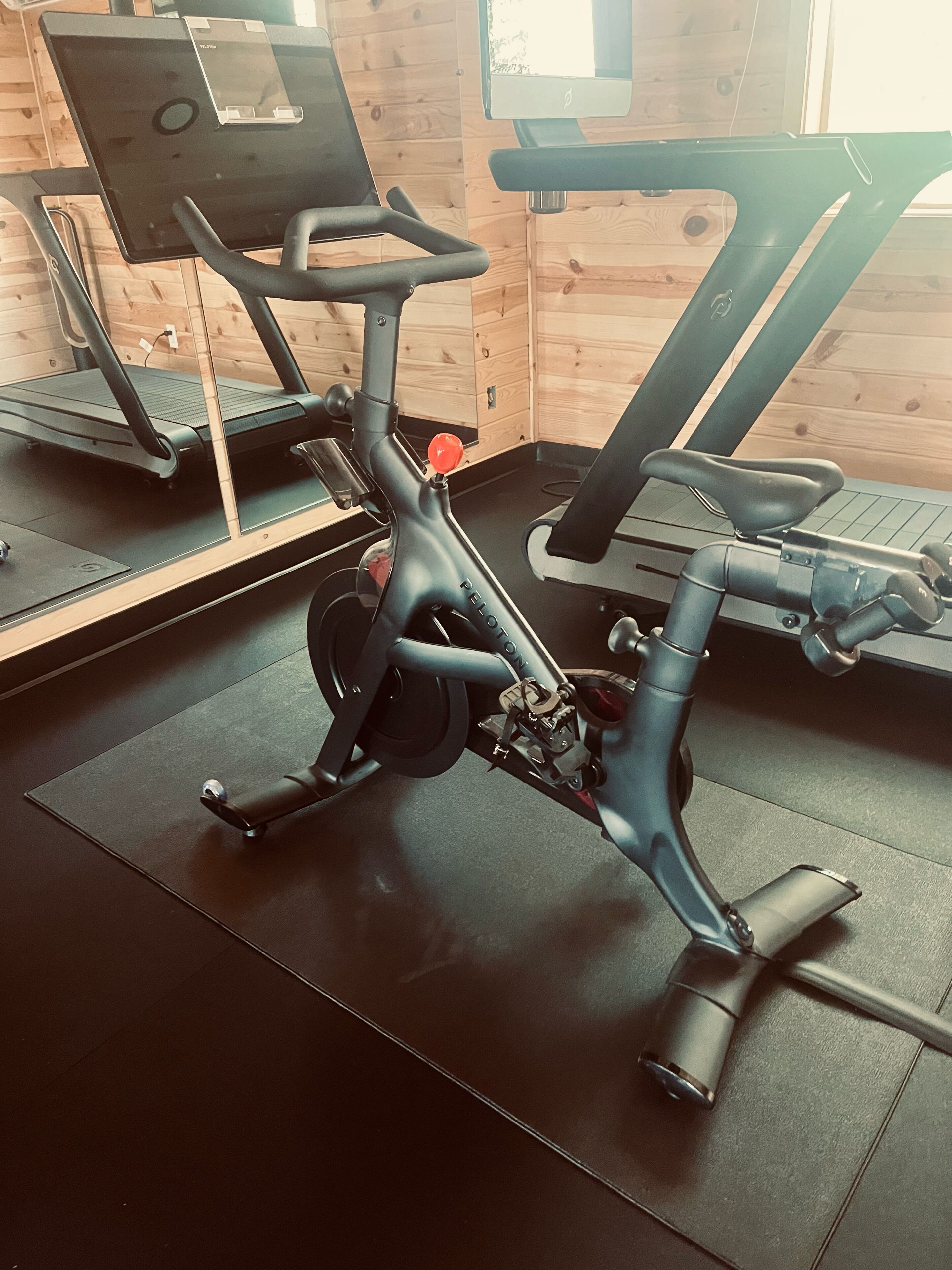Un vélo d’appartement, doté d’un écran numérique, est placé sur un tapis noir dans une pièce aux murs en bois et dotée d’une fenêtre.