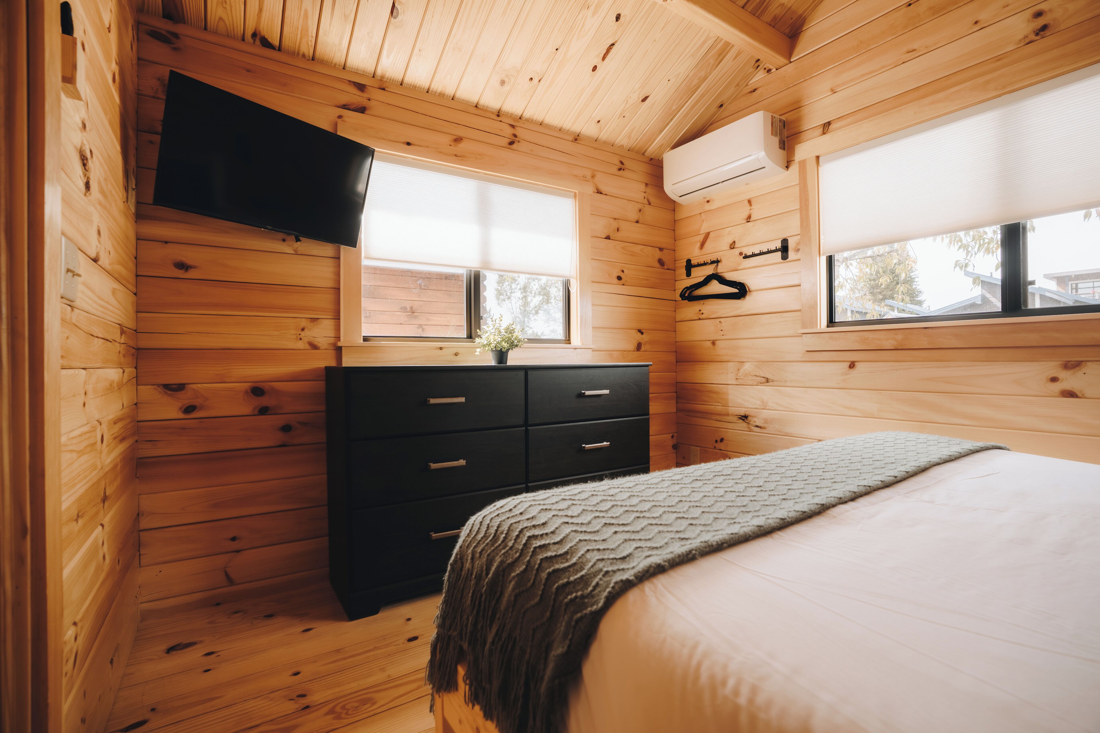 A wooden cabin bedroom with a bed, dresser, TV, and windows.