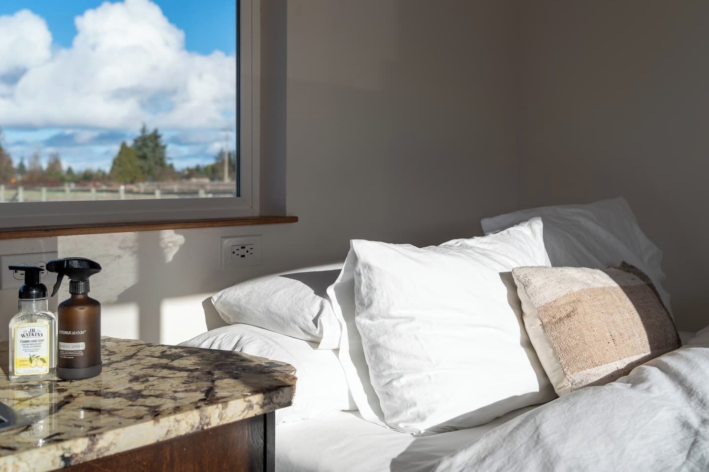 A bedroom with a bed, white pillows, a window with a view of trees and sky, and toiletries on a marble countertop.