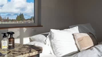 A bedroom with a bed, white pillows, a window with a view of trees and sky, and toiletries on a marble countertop.