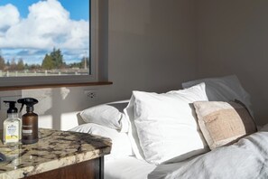Une chambre à coucher avec un lit, des oreillers blancs, une fenêtre donnant sur les arbres et le ciel, et des articles de toilette posés sur un comptoir en marbre.