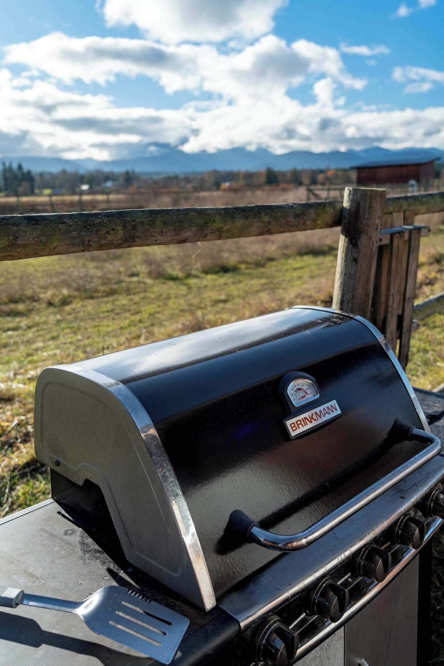 A black Bravkman grill with a digital thermometer, a spatula, and a handle, set against a rural backdrop with a wooden fence and a clear sky.