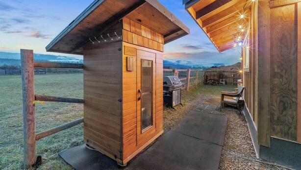 A wooden sauna structure with a grill outside, a covered seating area, and a view of the landscape. - Blue Moon Tiny House | Hot Tub · Sauna · Olympic Views (Port Angeles)