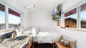 A compact bedroom with a bed, bedside table, and a view of a wooden cabin through a window. - Blue Moon Tiny House | Hot Tub · Sauna · Olympic Views (Port Angeles)