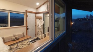 A compact bedroom with a large window, a built-in sink, and a bench with pillows. - Blue Moon Tiny House | Hot Tub · Sauna · Olympic Views (Port Angeles)