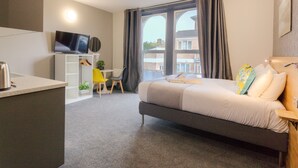 Un dormitorio con una cama, un escritorio, una silla, un televisor y vista a un edificio por la ventana.