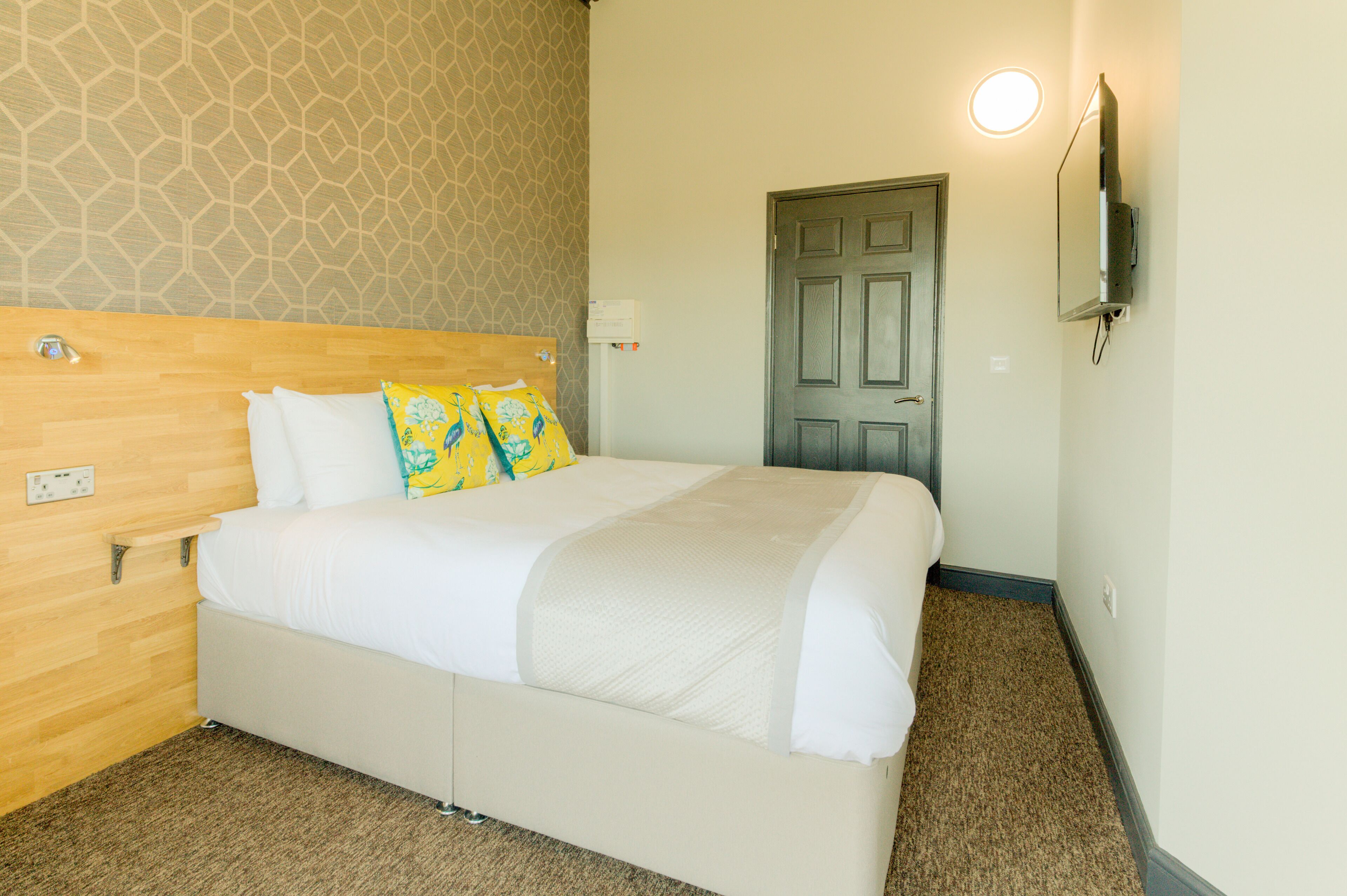A neatly made bed with white linens and two decorative pillows, a wooden headboard, a wall-mounted TV, and a door with a light above it.