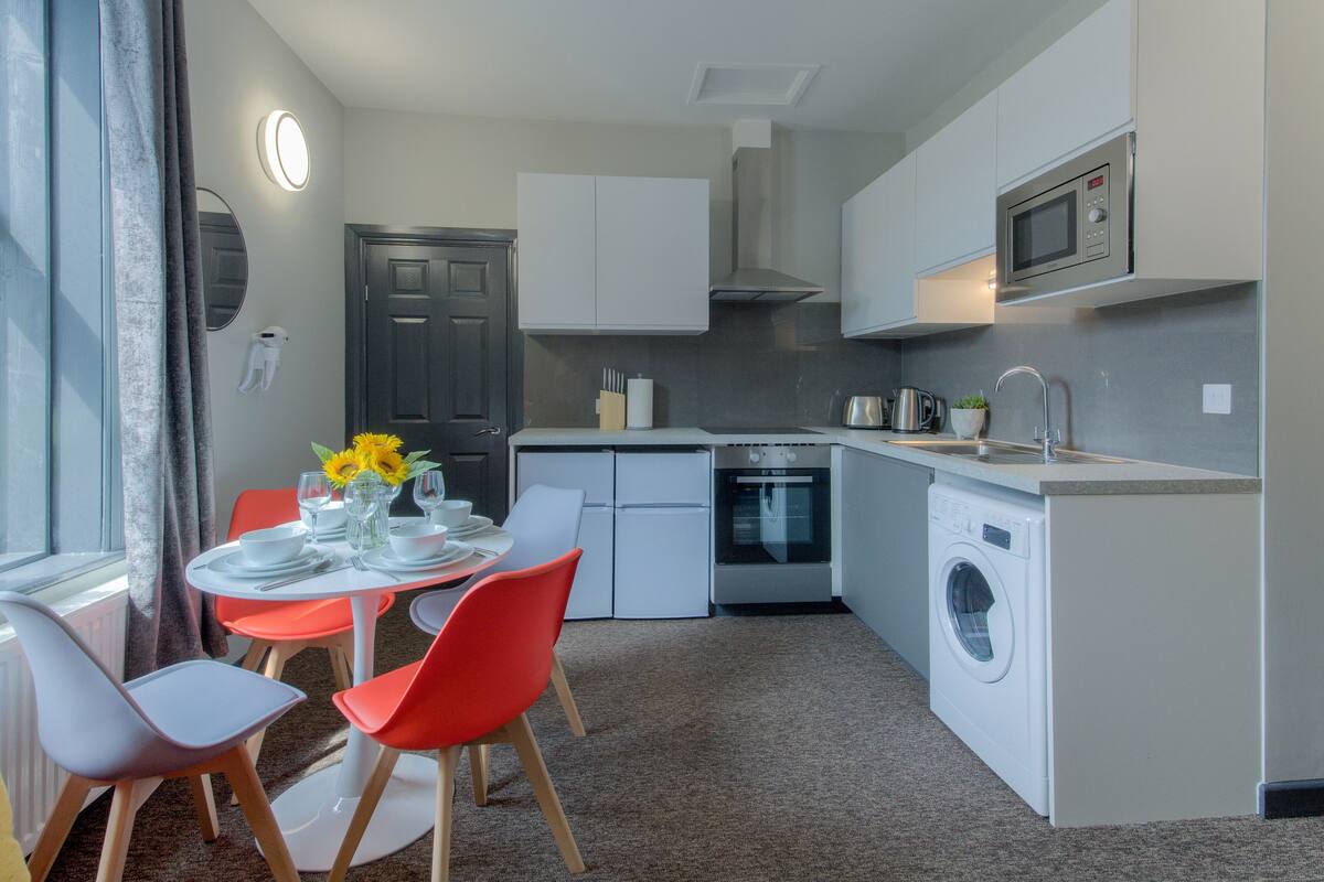 Une cuisine compacte équipée d’un four à micro-ondes, d’un four et d’un lave-linge. Elle comprend également un coin repas avec une table et des chaises.