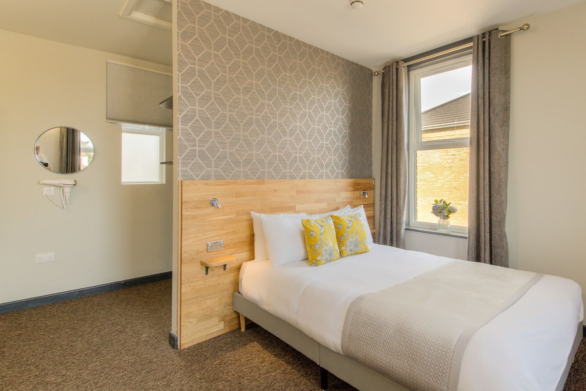 Une chambre d’hôtel avec une tête de lit en bois, un grand lit avec une literie blanche et des oreillers jaunes, un miroir rond et une fenêtre avec des rideaux.