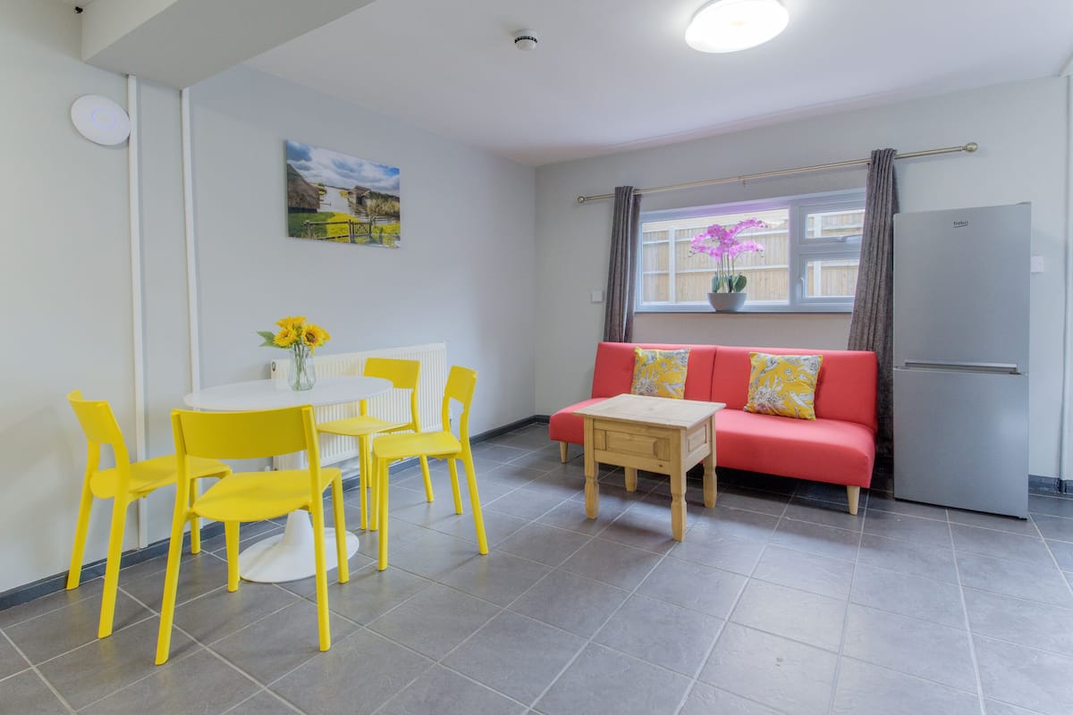 Une pièce comprenant une table à manger et des chaises, un canapé, une petite table en bois, un radiateur, une fenêtre avec des rideaux et un réfrigérateur.
