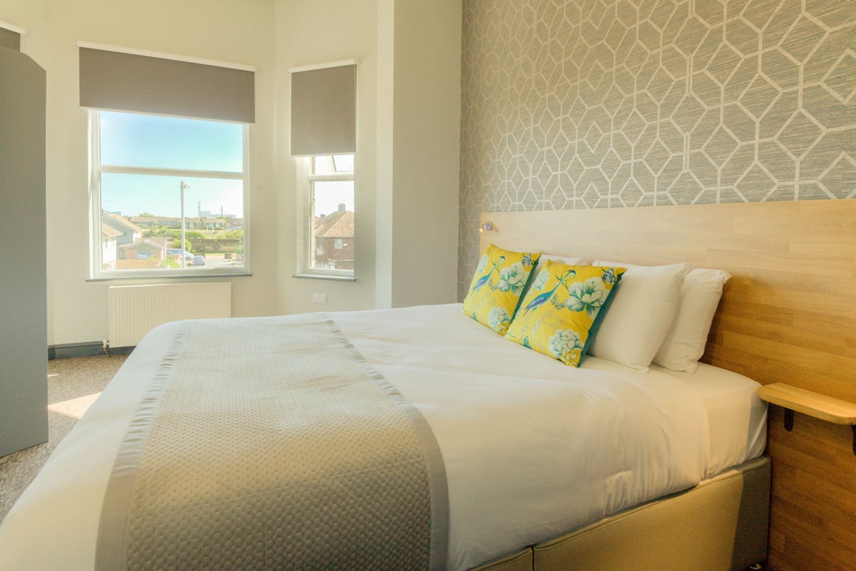 Une chambre d’hôtel avec un grand lit, une fenêtre donnant sur des immeubles et un mur orné d’un motif.