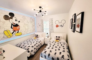 A children's room with two beds, Mickey Mouse wall art, and a ceiling light fixture.