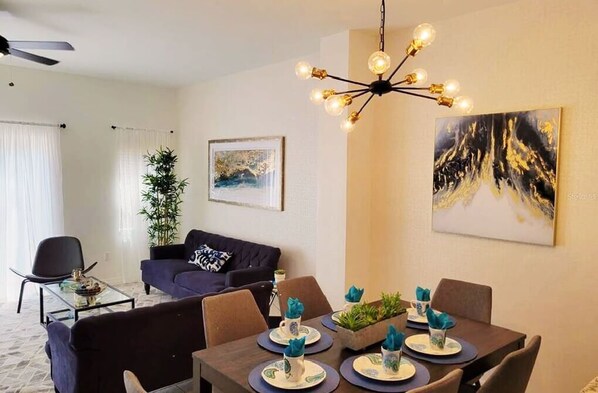 A modern living room with a dining area, featuring a ceiling fan, a chandelier, and artwork on the walls.