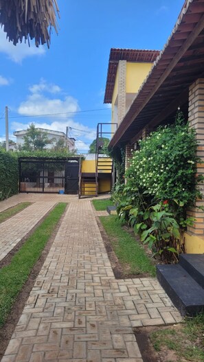 A paved pathway leading to a yellow building with a brick facade and a wooden roof. - Pousada Ello (São Miguel do Gostoso)