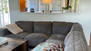 A grey sofa with a patterned throw blanket, a small table with a box, and a window with curtains and plants. - Dog 🐾 friendly home with a view 🌅 near airport ✈️ (Des Moines)