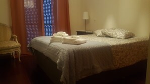 A neatly made bed with a patterned bedspread, a stack of folded towels, a bedside lamp, and a chair in a room with a window with blinds. - Holiday Apartment in Madeira (Caniço)