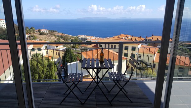 A balcony with a view of the sea, a bottle of liquor, and a wooden table with chairs. - Holiday Apartment in Madeira (Caniço)