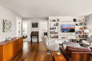 A modern living room with a wooden floor, a grey armchair, a brown leather sofa, a wooden cabinet, and a bookshelf filled with books and a TV displaying a scenic view.