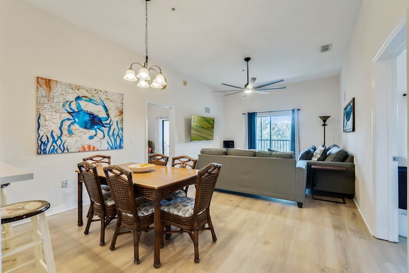 Zona pranzo con tavolo in legno e sedie in vimini, ventilatore a soffitto e un grande quadro appeso al muro.