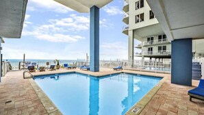 A pool area with a clear view of the ocean, lounge chairs, and a multi-story building with balconies.