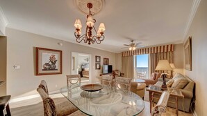 A spacious living room with a glass-top dining table surrounded by patterned chairs, a chandelier, a ceiling fan, and a large window with curtains.