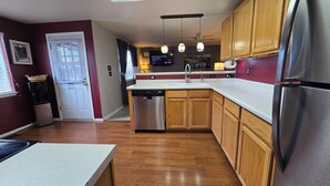 A kitchen with wooden cabinets, a stainless steel refrigerator, a dishwasher, and a built-in oven. There is a window with blinds, a wall-mounted TV, and pendant lights above the sink. - Cozy Colorado home with Serene views meant for pure relaxation! (Henderson)