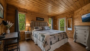 A wooden cabin bedroom with a bed, bedside tables, a dresser, a desk with a lamp, and a TV on the wall. - Ranked Top 1% Cabin-Location-Views-Hot Tub-Games (Pigeon Forge)