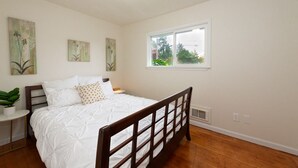 Un dormitorio con cama de madera, ropa de cama blanca, una mesita de noche con una planta y una ventana con vistas a la vegetación.