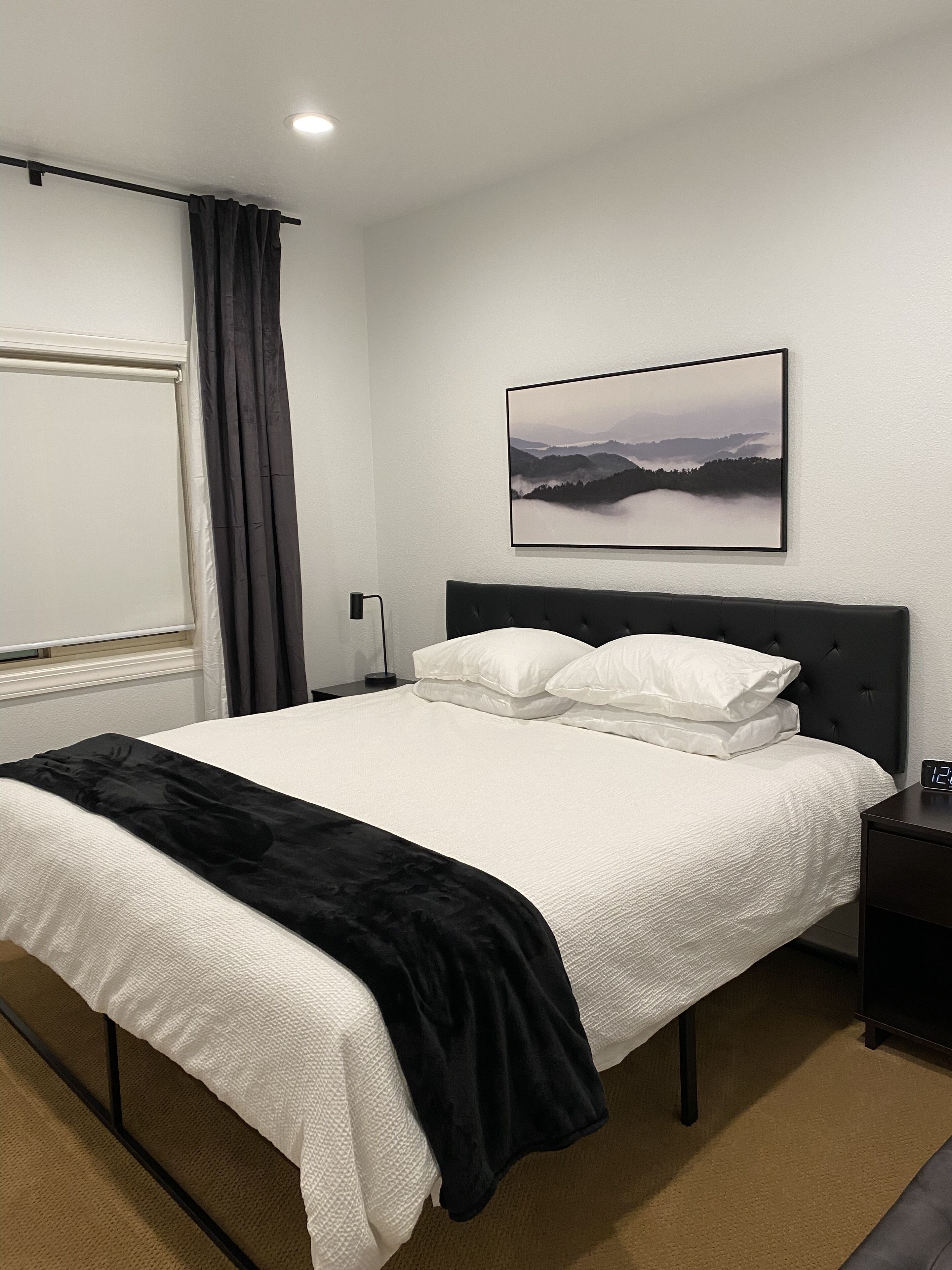 Una cama bien hecha con ropa blanca y un plaid negro, una mesita de noche con un reloj, y un cuadro enmarcado de un paisaje en la pared.