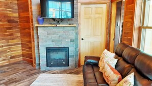 A cozy living room with a fireplace, a leather sofa, and a television mounted on the wall. - 2-bedroom vacation home near Mark Twain National Forest (Potosi)