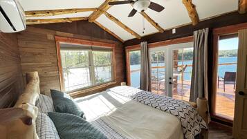 A bedroom with a large bed, wooden ceiling beams, a ceiling fan, and a view of a lake through large windows.