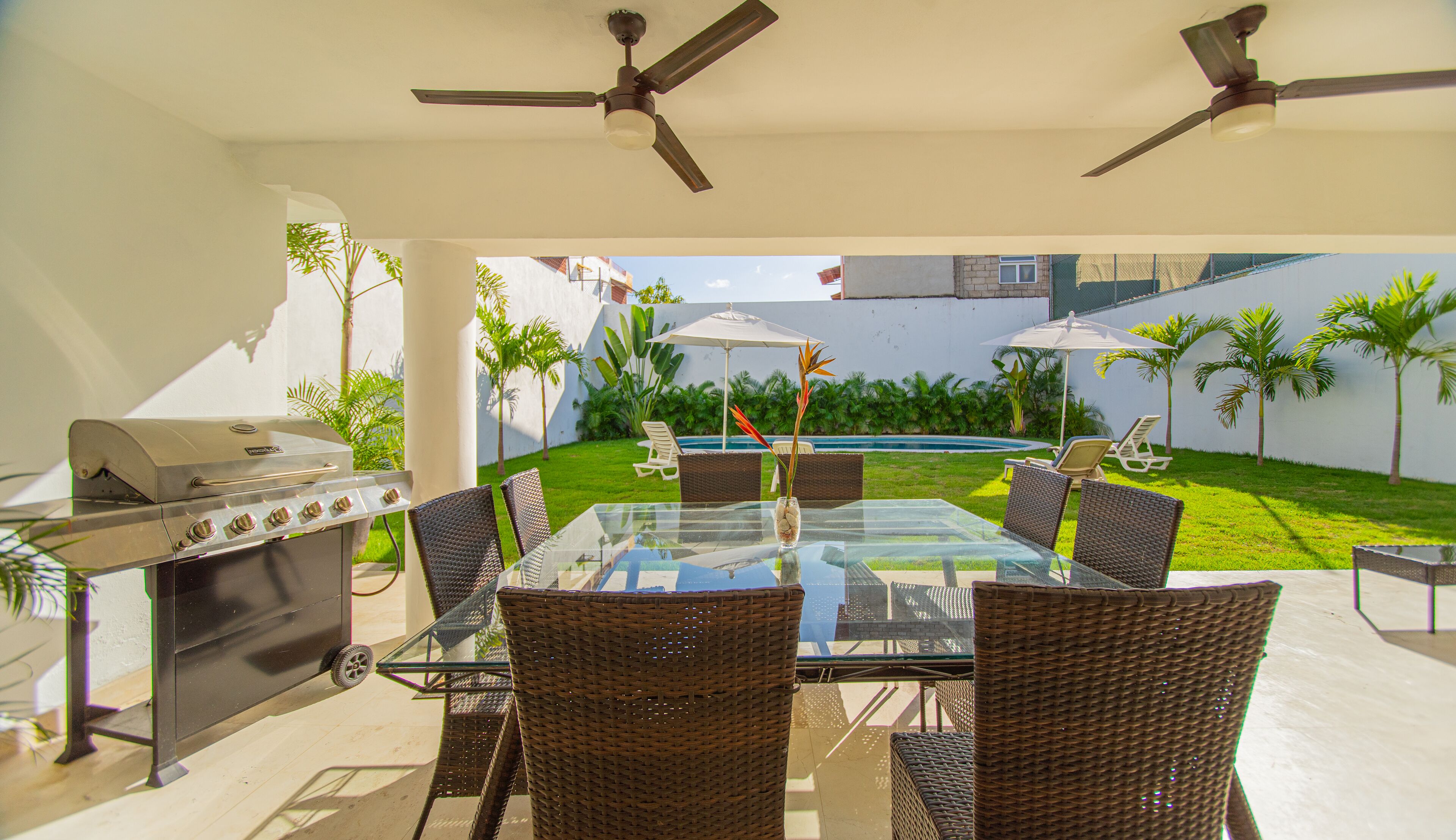 Eine Außenpatio mit Glastisch, Korbstühlen, Grill, Deckenventilatoren und Blick auf einen Swimmingpool und Grünflächen.