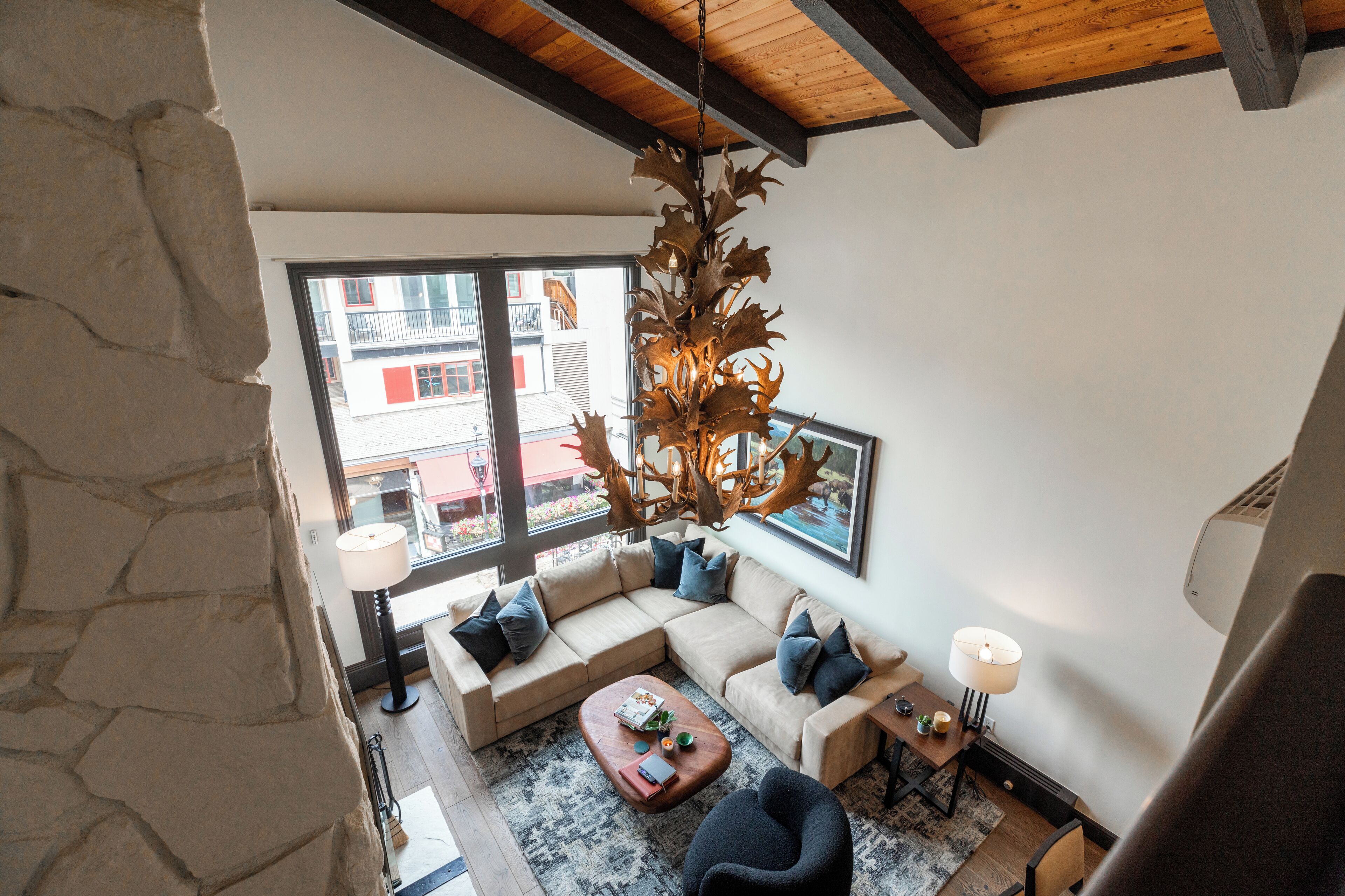 A modern living room with a stone wall, a large window, a wooden ceiling, a beige sectional sofa, a coffee table, and a unique chandelier.
