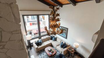 A modern living room with a stone wall, a large window, a wooden ceiling, a beige sectional sofa, a coffee table, and a unique chandelier.