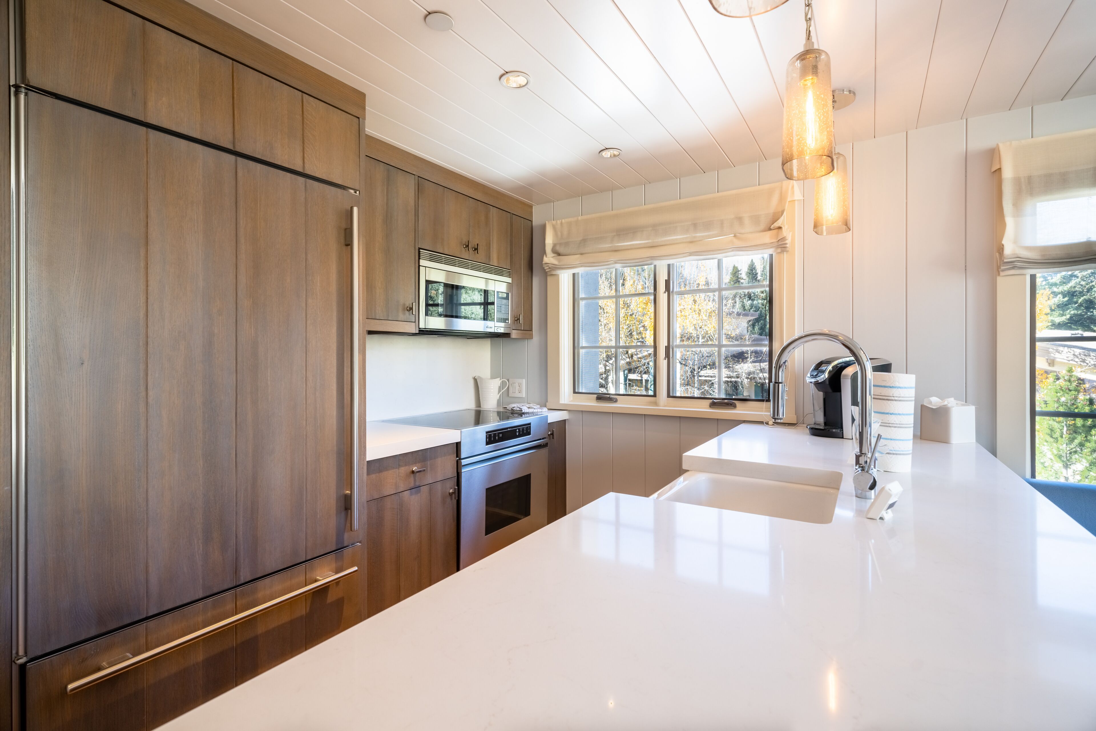 A modern kitchen with wooden cabinets, stainless steel appliances, and a large window providing a view of trees.