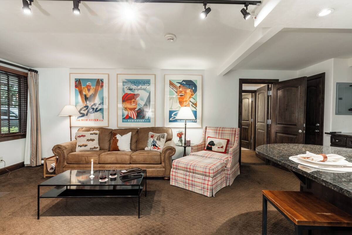 A living room with a sofa, coffee table, and ottoman, a kitchen area with a granite countertop, and framed sports posters on the wall.