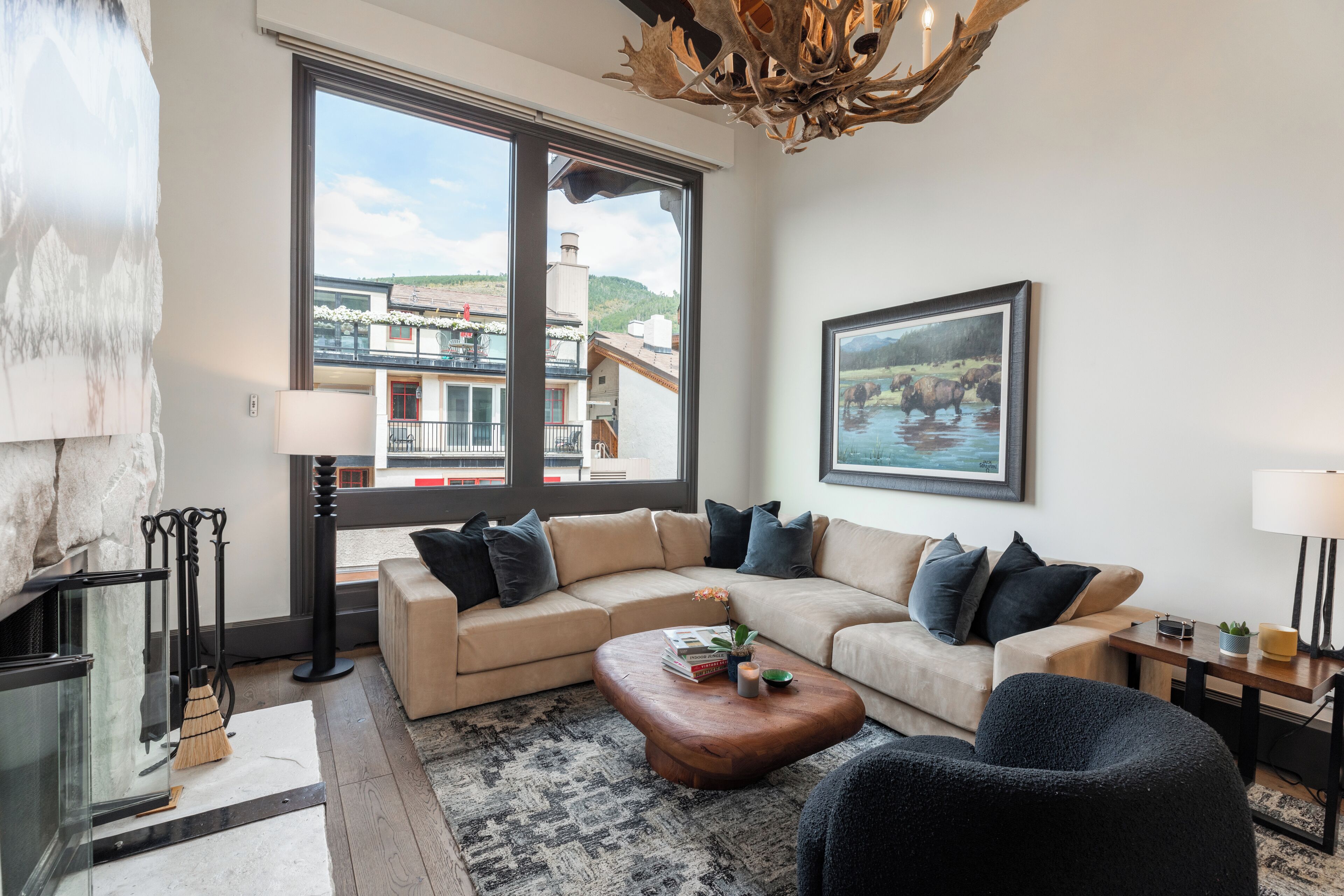 A modern living room with a beige sectional sofa, a wooden coffee table, and a large window offering a view of buildings and hills.