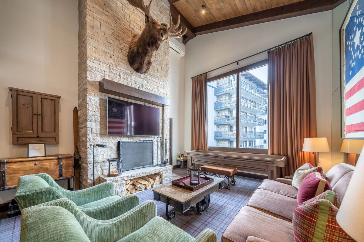 A cozy living room with a stone fireplace, a mounted deer head, a flat-screen TV, comfortable seating, and a large window with a view of a building.