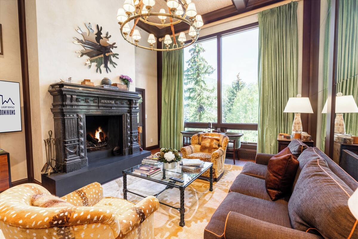 A cozy living room with a fireplace, comfortable seating, and a large window offering a view of greenery.
