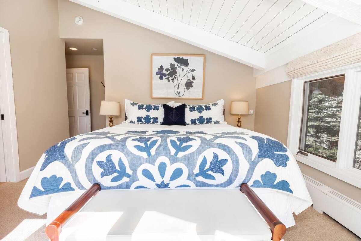 A bedroom with a large bed, two bedside lamps, a framed floral artwork above the bed, and a window with a view of trees.